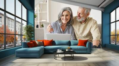 Happy mature mid age couple using laptop sit on sofa doing ecommerce shopping online on website. Smiling senior old adult man and woman looking at computer buying insurance browsing internet at home. Wall mural