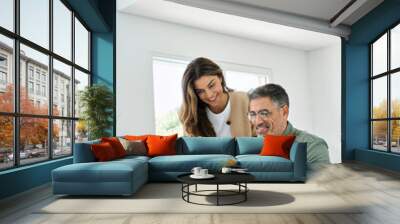 Happy mature older couple using laptop technology at home. Smiling middle aged senior man working on computer sitting at table with wife standing nearby in living room. Vertical candid shot. Wall mural