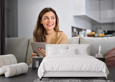 Happy mature older woman using digital tablet sitting on couch at home. Smiling middle aged 45 years old woman looking away at copy space holding tab computer relaxing on sofa in living room. Wall mural