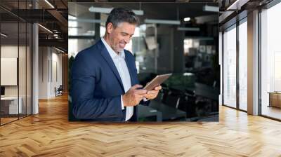 Happy mid aged business man ceo wearing blue suit standing in office using digital tablet. Mature businessman professional executive manager holding tab working on financial data on technology device. Wall mural