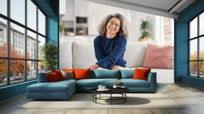 Happy middle aged woman in her 50s sitting on sofa in living room at home. Smiling beautiful mature older lady with curly gray hair relaxing on couch looking at camera on couch in cozy house. Portrait Wall mural