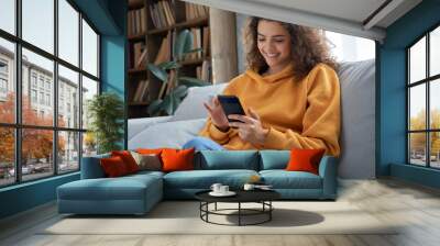 Happy millennial hispanic teen girl checking social media holding smartphone at home. Smiling young latin woman using mobile phone app playing game, shopping online, ordering delivery relax on sofa. Wall mural