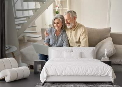 Happy old mid age family couple using laptop computer sitting on couch. Smiling senior mature husband and wife doing ecommerce online shopping, browsing web, watching tv, having video call at home. Wall mural