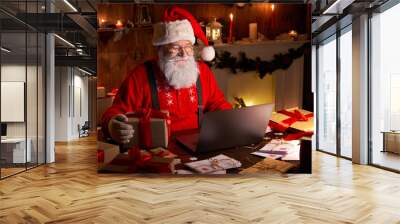 Happy old Santa Claus wearing hat holding gift box using laptop computer sitting at workshop home table late on Merry Christmas eve. Ecommerce website xmas time holiday online shopping e commerce sale Wall mural