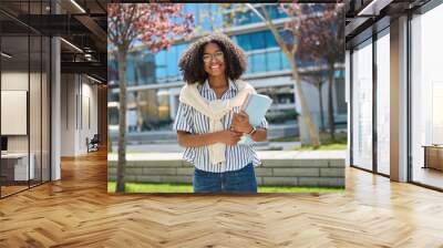 Happy pretty female African American student walking outside university. Smiling cheerful girl model with curly hair wearing glasses, holding notebooks looking at camera outdoors, portrait. Wall mural