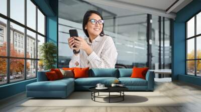 Happy professional Asian business woman using phone looking away in office. Young smiling female manager holding cellphone looking away thinking on new cell application sitting at desk in office. Wall mural