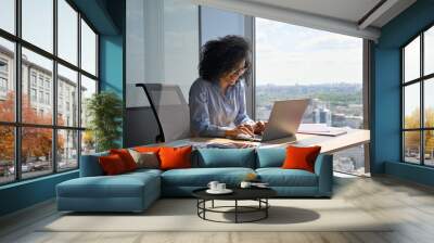 Happy smiling African American businesswoman executive manager sitting at desk working typing on laptop computer in contemporary corporation office with panoramic view. Business technologies concept. Wall mural