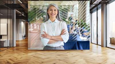 Happy smiling confident middle aged Asian older senior female leader businesswoman standing in modern office workplace looking at camera arms crossed. Business successful executive concept. Portrait. Wall mural