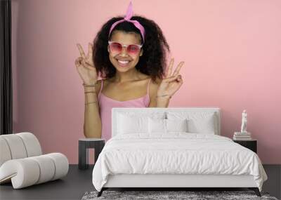 Happy smiling fashion young african teen hipster girl black woman wearing stylish glasses looking at camera show peace hand sign isolated on light pink summer studio background, portrait, copy space Wall mural