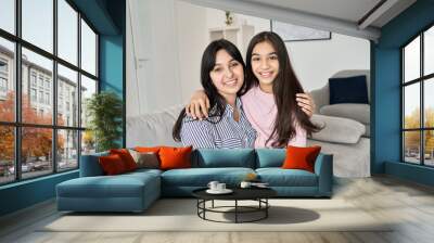 Happy teen child daughter hugging young indian mother at home, portrait. Smiling ethnic family mom with teenage kid girl embracing, bonding together looking at camera sitting on couch. Wall mural