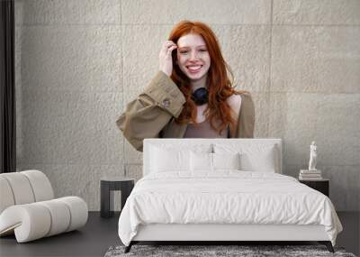 Happy teen stylish cool redhead fashion girl model standing on urban wall background looking at camera. Portrait of beautiful smiling teenage girl with red hair wearing trendy clothes laughing outdoor Wall mural