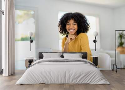 Happy young African American lady sitting at home table and looking at camera while hybrid working or elearning online on laptop, smiling woman with computer in living room. Portrait. Wall mural