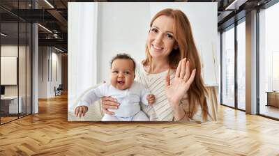 Happy young caucasian mother waving hand holding little cute funny african american baby girl daughter on lap having virtual distance meeting video call looking at camera at home, webcam view. Wall mural