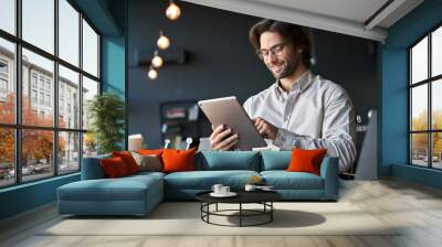 Happy young European business man entrepreneur holding tad computer at work. Male professional employee worker using digital tablet fintech device sitting in office checking financial online data. Wall mural