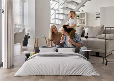 Happy young family couple having fun playing with happy cute kid boy sitting on dads shoulders in modern living room. Parents and toddler child son enjoying spending time sitting on floor at home. Wall mural