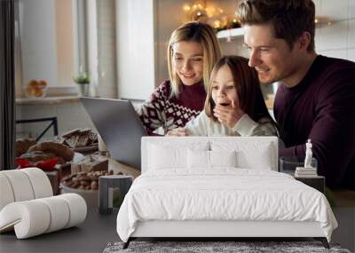 Happy young parents with cute excited small kid daughter using laptop computer at home sitting at Christmas table having virtual party on video call, doing ecommerce family online shopping together. Wall mural