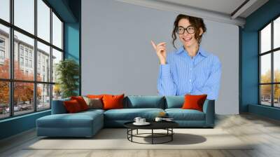 Happy young smiling professional business woman wearing blue shirt glasses looking away pointing finger at copy space showing aside presenting ads promotion standing isolated at gray background. Wall mural