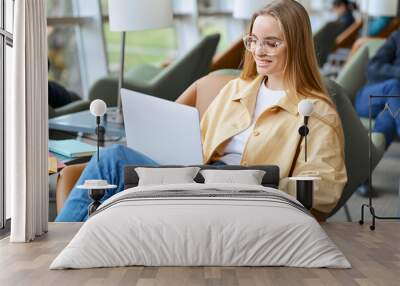 Happy young woman, smiling girl student wearing eyeglasses using laptop computer sitting on chair in university campus, coworking office or airport lounge space hybrid learning or working online. Wall mural