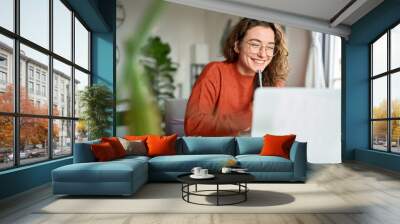 Happy young woman using laptop sitting at desk writing notes while watching webinar, studying online, looking at pc screen learning web classes or having virtual call meeting remote working from home. Wall mural