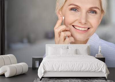 Headshot portrait of attractive senior adult older blonde lady wears bathrobe in bathroom applying nourishing tightening lifting face skin care cream treatment, looking at camera. Wall mural