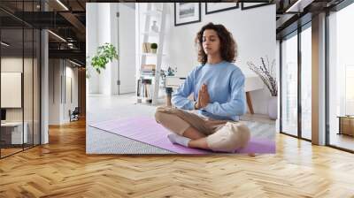 Healthy serene young woman meditating at home with eyes closed doing pilates breathing exercises, relaxing body and mind sitting on floor in living room. Mental health and meditation for no stress. Wall mural