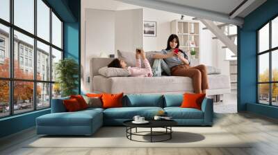 Indian family mother with teen daughter using phones devices at home. Young mom with teenage child holding cellphones relaxing together on couch in living room. Social media technology addiction. Wall mural