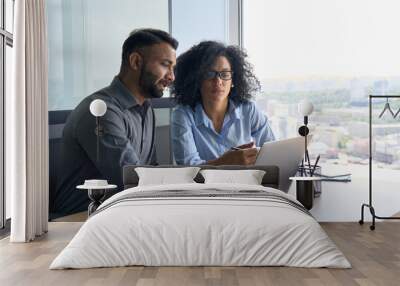 Indian male ceo executive manager mentor giving consultation on financial operations to female African American colleague intern using laptop sitting in modern office near panoramic window. Wall mural