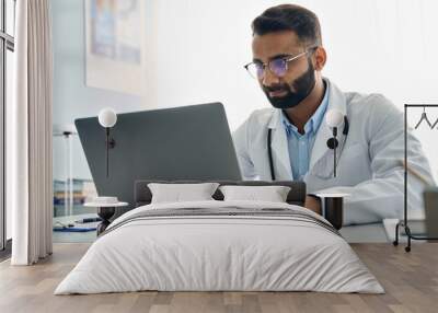 Indian male doctor medic expert wearing white coat using laptop computer at work in hospital, checking patients electronic files medical forms, having online virtual telehealth consultation. Wall mural