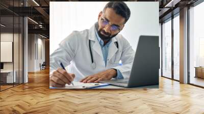 Indian male medical worker in modern clinic wearing eyeglasses and white doctor's coat using laptop computer writing health personal data, consulting remotely. Telemedicine healthcare concept. Wall mural