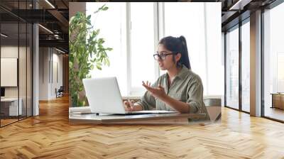 Indian woman online tutor remote teacher wearing glasses speaking to webcam chat explaining online class zoom video call school lesson looking at laptop virtual conference meeting work at home office. Wall mural