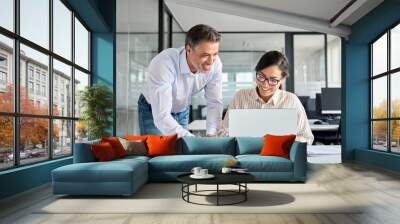 Mid aged Latin manager mentor helping Asian worker working on laptop in office. Two happy professional coworkers discussing online plan at work. Diverse business people using computer and talking. Wall mural