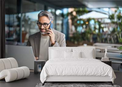 Middle aged business man talking on mobile phone hybrid working on laptop sitting outside office. Mature businessman making call on cellphone speaking to client using computer outdoors. Copy space. Wall mural