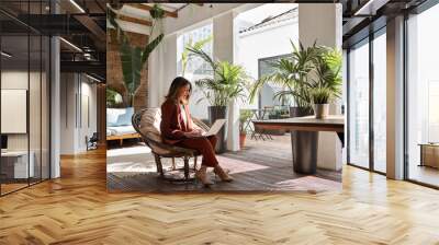 Middle aged business woman using laptop sitting in chair in sunny green office. Busy professional mature female entrepreneur or executive wearing suit holding computer on lap working. Candid photo. Wall mural