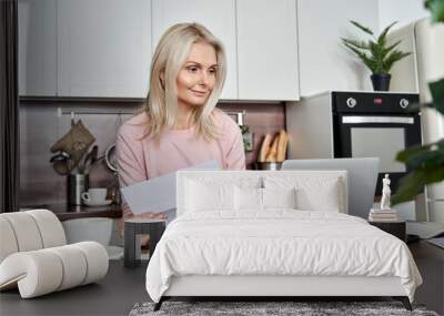 Middle aged mature woman holding paper bill or letter using laptop computer at home for making online payments on website, calculating financial taxes fee cost, reviewing bank account, loan rates. Wall mural