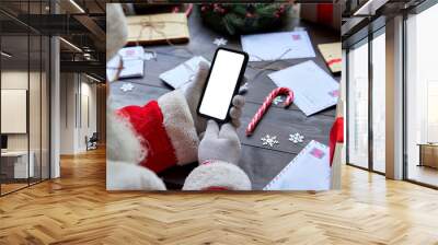 Over shoulder view of Santa Claus wearing costume holding smartphone with white blank display screen using mobile app sitting at workshop table on Merry Christmas eve. Ecommerce application ad closeup Wall mural