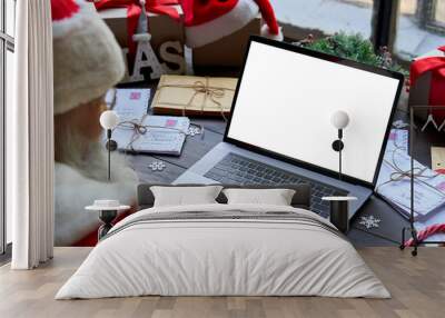 Over shoulder view of Santa Claus wearing costume using laptop computer with white blank empty mock up screen monitor sitting at workshop table on Merry Christmas eve. E commerce website ads concept Wall mural