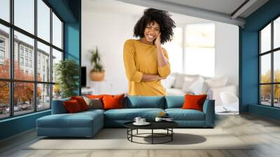 Portrait of smiling happy young pretty 25 years old African American woman wearing yellow jumper and jeans standing in modern living room looking at camera in modern cozy home. Wall mural