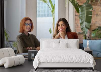 Portrait of two happy business women of young and middle age entrepreneurs or employees working together sitting at work desk with laptop computer looking at camera in modern cozy green office. Wall mural