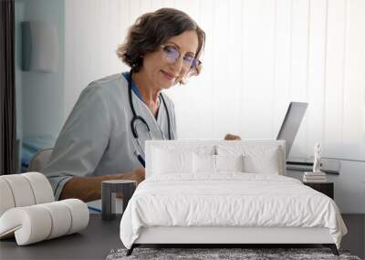 Senior middle aged female medical worker in modern clinic wearing white doctor's coat having videocall using laptop writing health personal data, consulting remotely. Telemedicine healthcare concept. Wall mural