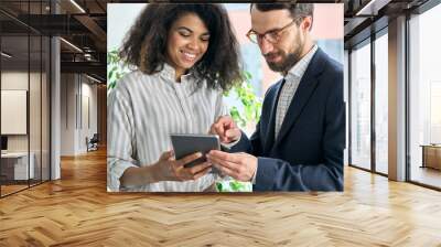 Serious executive middle aged ceo bearded boss businessman holding using looking at tablet device and happy smiling young African American female manager standing discussing project report at office. Wall mural