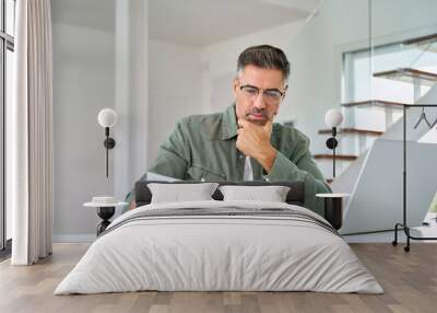 Serious middle aged mature man entrepreneur wearing eyeglasses looking at paper bill checking insurance invoice or tax document calculating expenses working at home using laptop computer. Wall mural