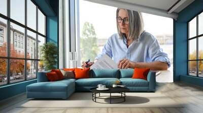 Serious older lawyer businesswoman in glasses reading signing trust partnership contract sit at table in office. Executive ceo put signature make legal bank sale financial loan investment agreement. Wall mural