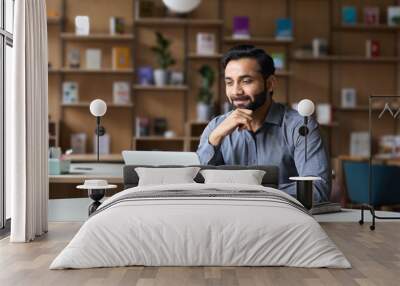 Smiling bearded indian businessman working on laptop in modern office lobby space. Young indian student using computer remote studying, watching online webinar, virtual training on video call meeting. Wall mural