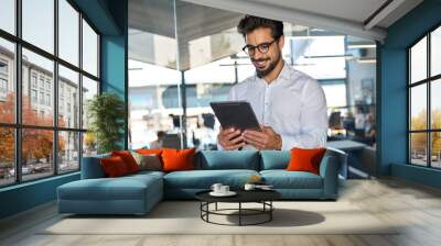 Smiling busy young Latin business man entrepreneur using tablet standing in office at work. Happy male professional executive manager using tab computer managing financial banking or marketing data. Wall mural