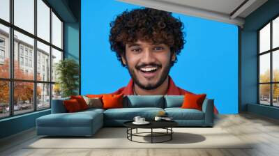 Smiling cheerful young curly bearded indian cool positive guy laughing isolated on blue background. Happy ethnic man looking at camera having fun posing for headshot close up face portrait. Wall mural