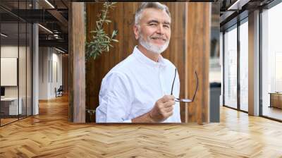 Smiling classy older senior business man looking at camera standing outdoors. Happy successful 60 years old businessman wearing white shirt holding glasses posing for portrait outside. Wall mural