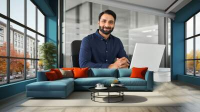 Smiling confident Indian business man employee looking at camera sitting at work desk with laptop computer. Portrait of smart happy businessman office worker or entrepreneur posing at modern workplace Wall mural