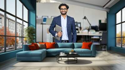 Smiling elegant young indian business man professional manager, arab company employee, eastern businessman executive financial banker wearing suit standing in modern office holding laptop, portrait. Wall mural