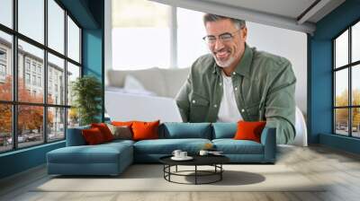 Smiling happy older mature middle aged man wearing eyeglasses looking at computer sitting at table, using laptop hybrid working online, elearning, browsing web, searching online at home. Vertical. Wall mural