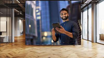 Smiling happy young eastern indian business man professional manager standing outdoor on street holding using digital tablet online fintech in night city with urban lights looking at camera, portrait. Wall mural